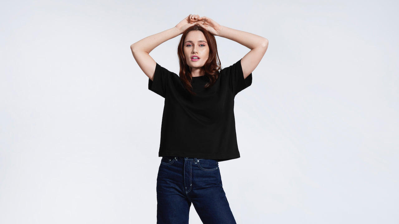 A woman with her hands on her head looking at the camera, modelling a plain black boxy, heavyweight organic cotton t shirt.