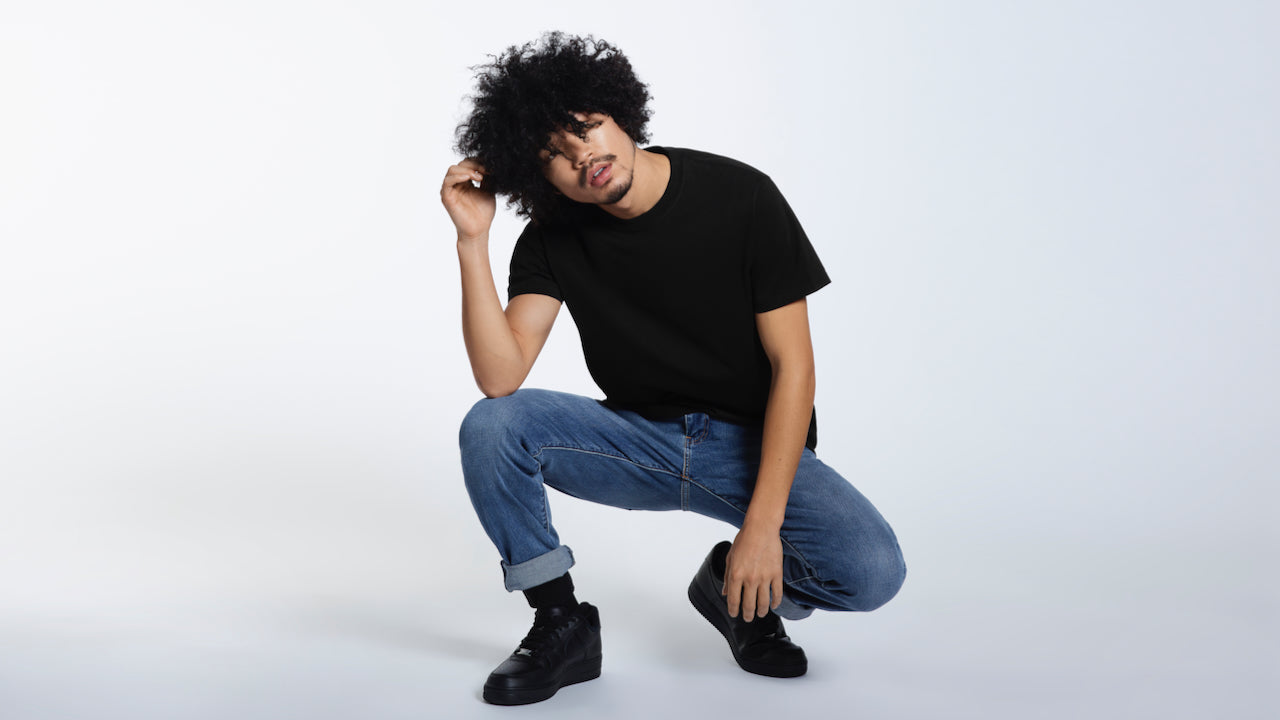 A man crouching down touching his hair, modelling a plain black organic cotton t shirt.