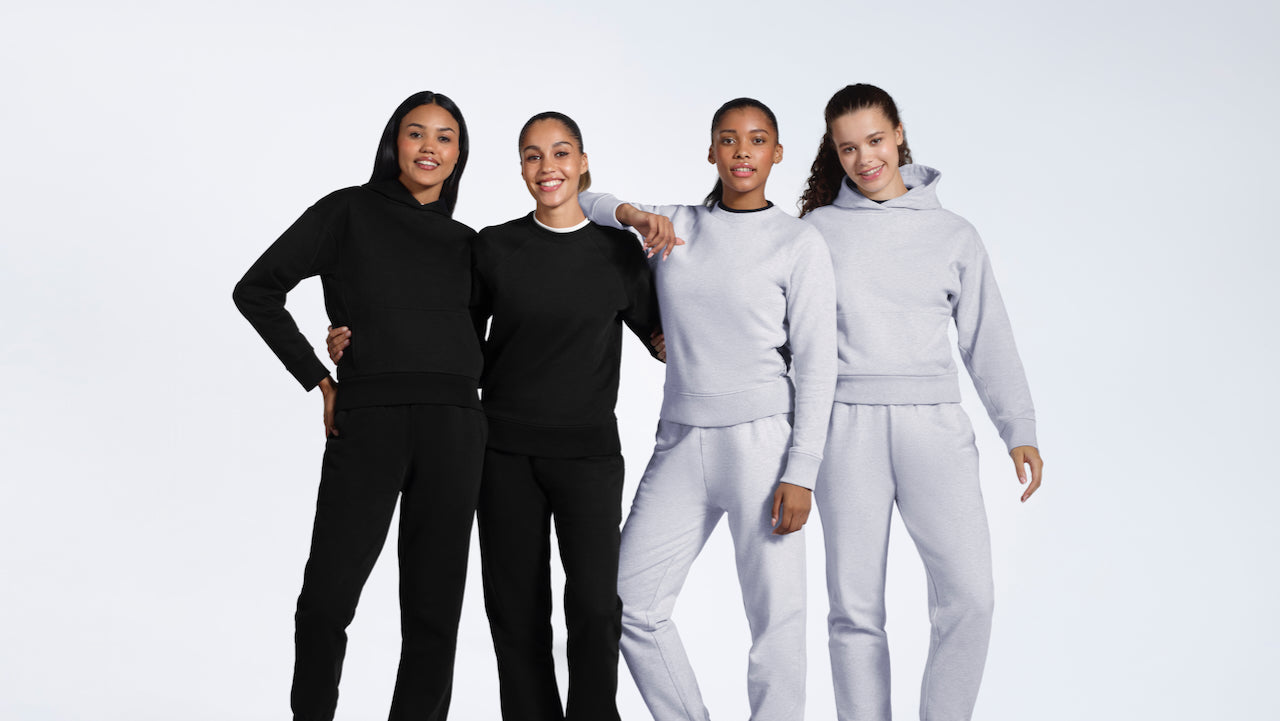 Four women smiling, modelling matching black and grey heavyweight organic cotton tracksuit sets.