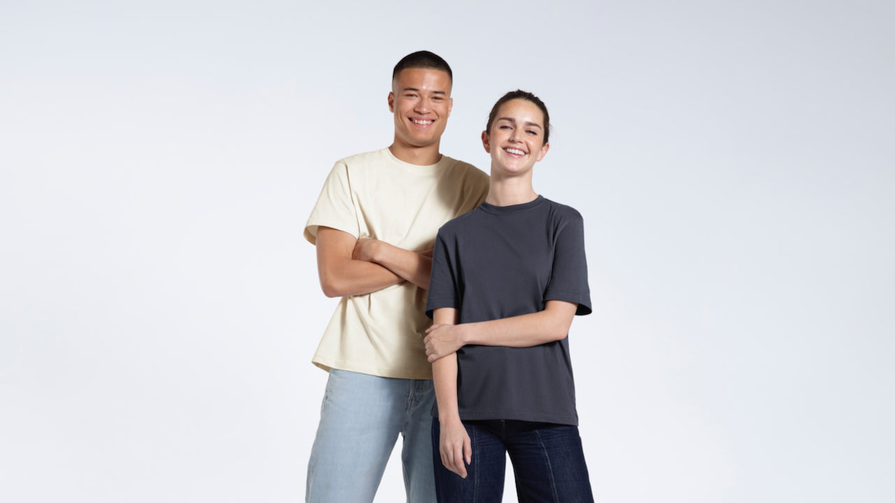 A man and woman looking into the camera smiling, modelling plain ecru and dark grey organic cotton t shirts.