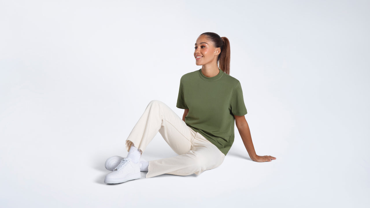 A woman sitting on the floor smiling, modelling a plain khaki green short sleeve organic cotton t shirt.