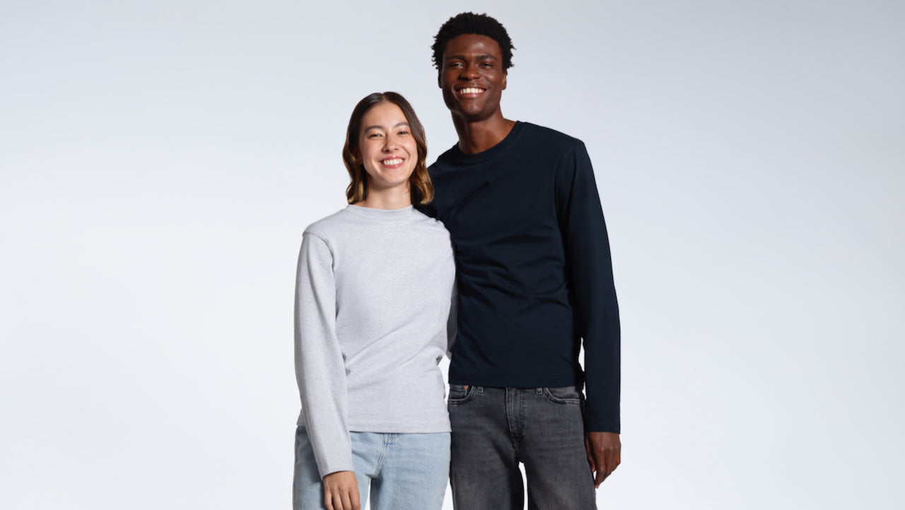 A man and woman smiling into the camera with their arms round eachother, modelling plain grey and navy organic cotton long sleeve t shirts.