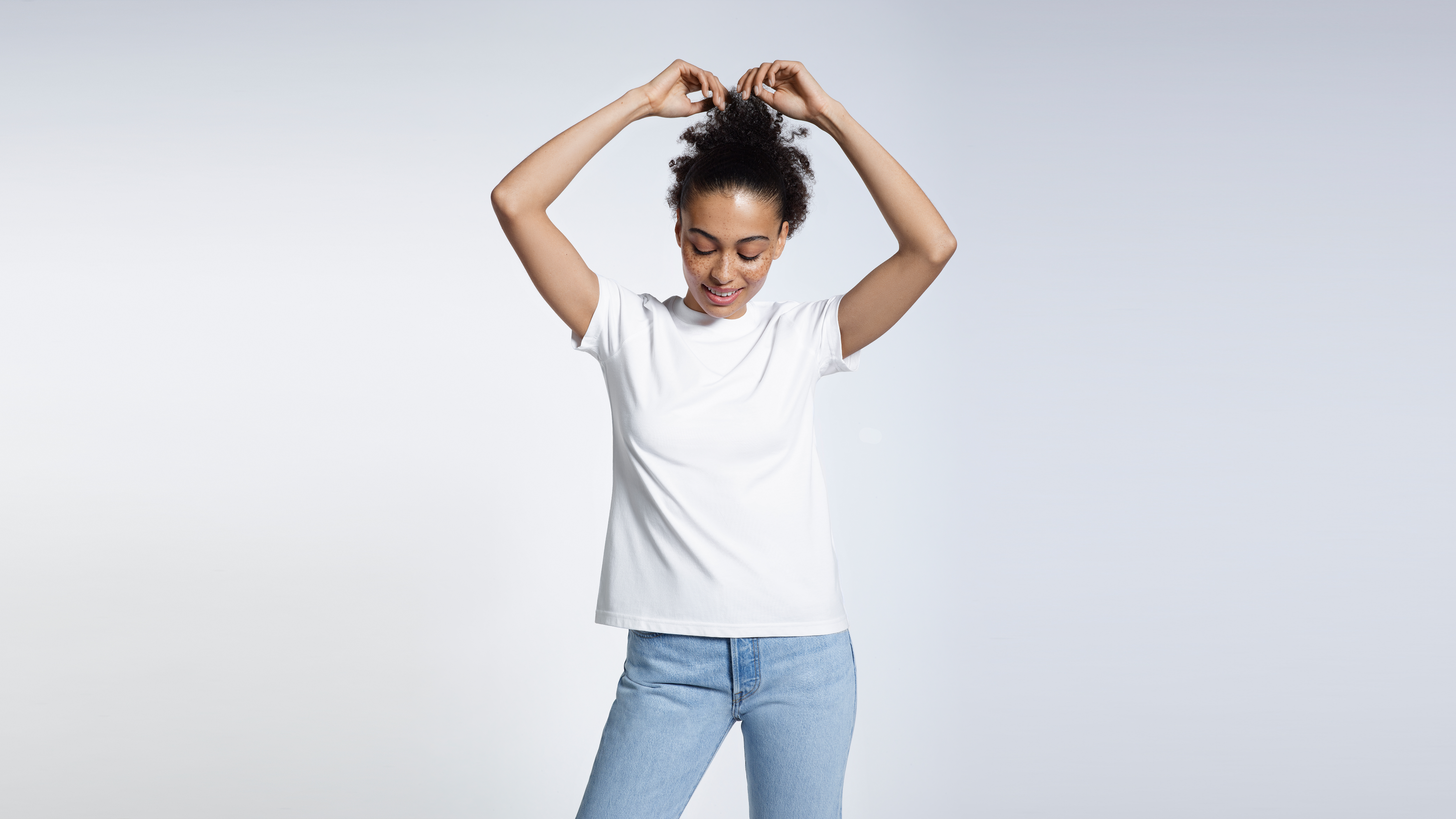 A woman with her arms above her head touching her hair, modelling a plain white short sleeve organic cotton t shirt.