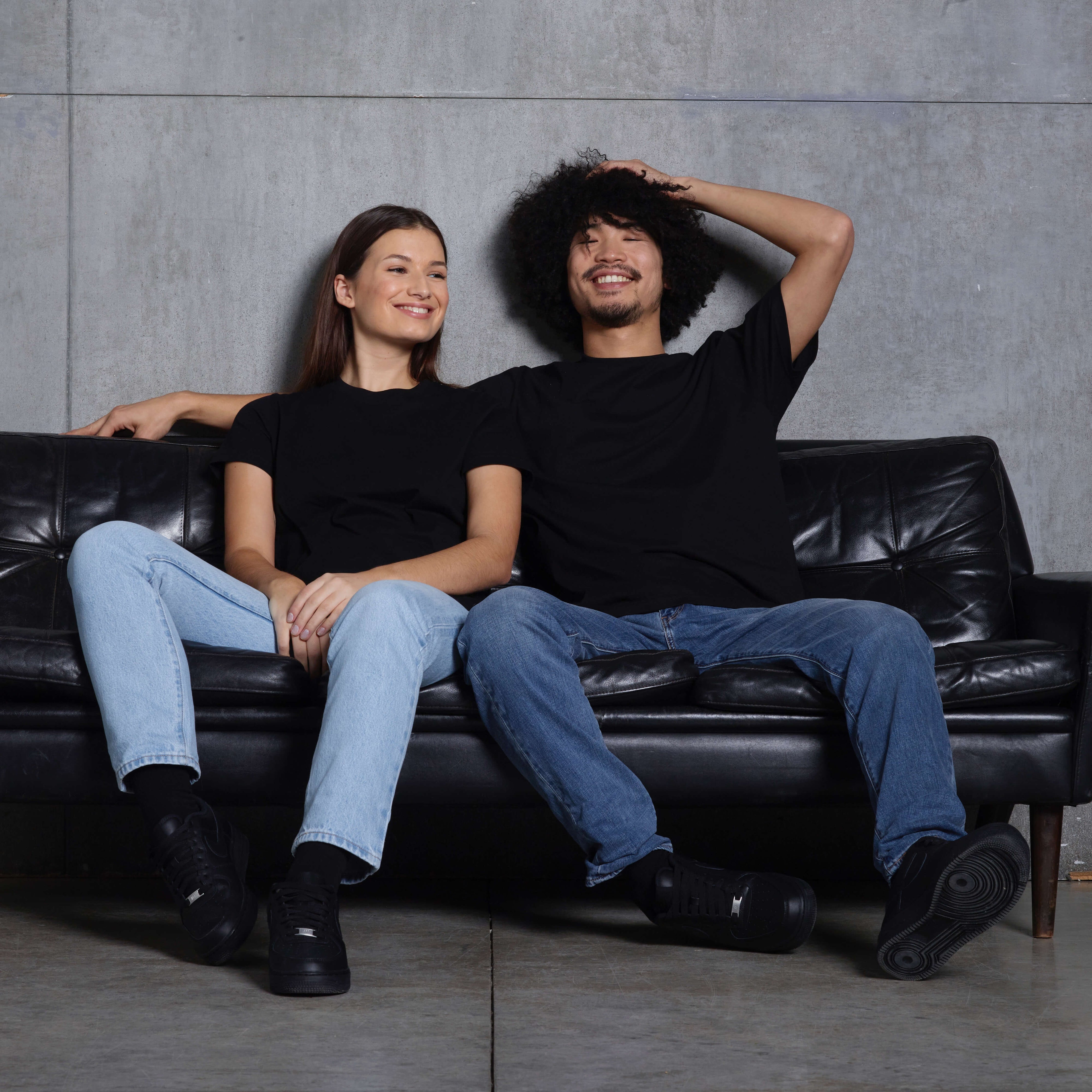 A man and woman sitting on a leather sofa, smiling, wearing plain black short sleeve organic cotton t shirts.