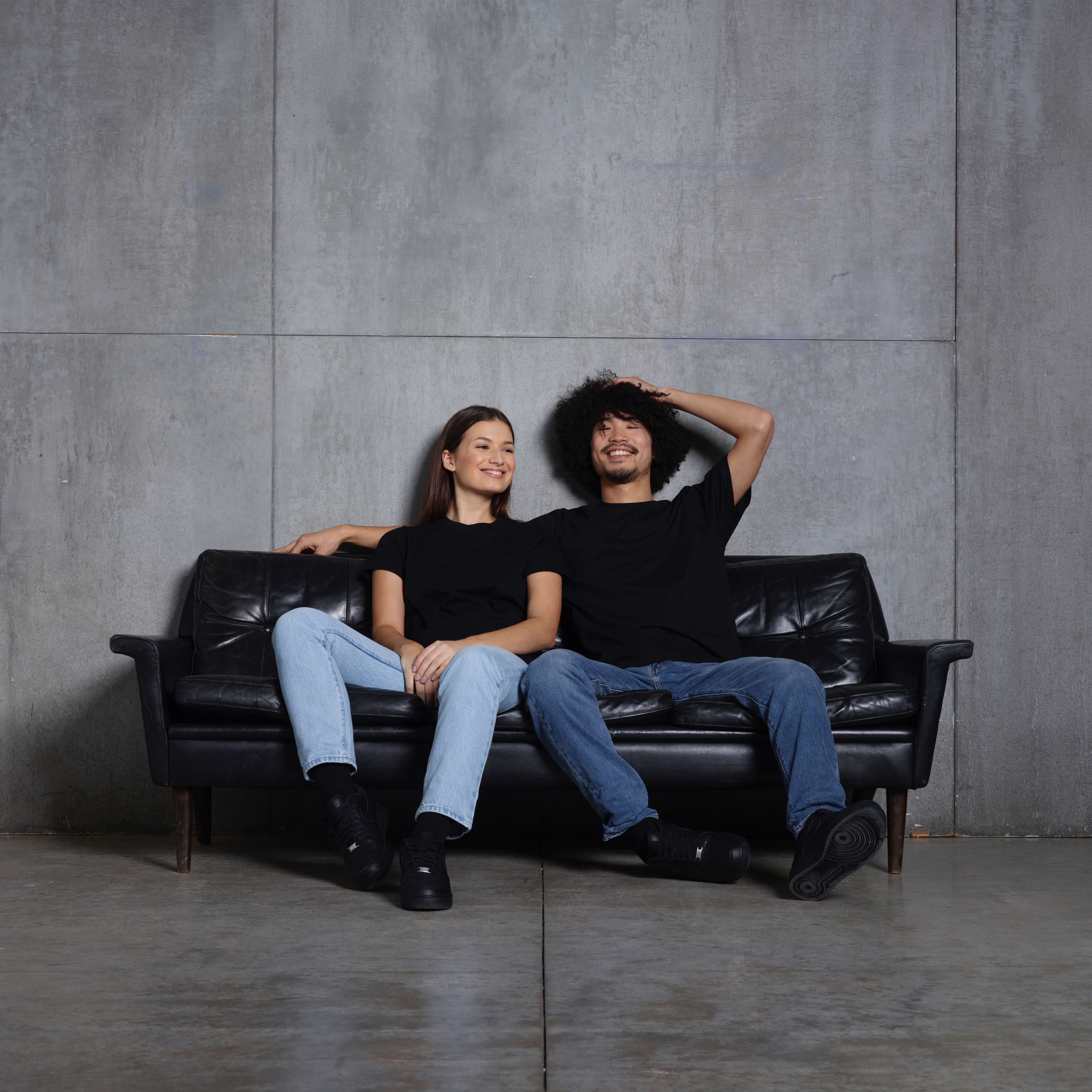 A man and woman sitting on a leather sofa, smiling, both wearing plain black t-shirts.