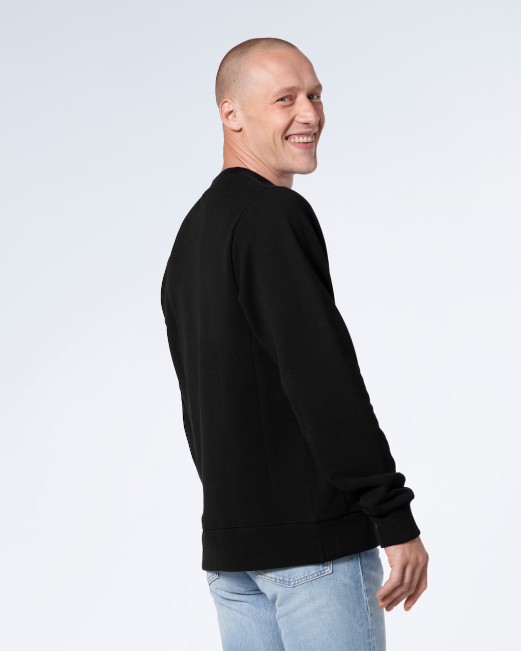 A man looking over his shoulder smiling at the camera, showing the back of his plain black french terry organic cotton sweatshirt.