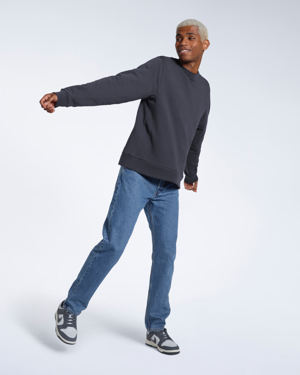 A full body photo of a man smiling looking away from the camera, modelling a plain dark grey french terry organic cotton sweatshirt.