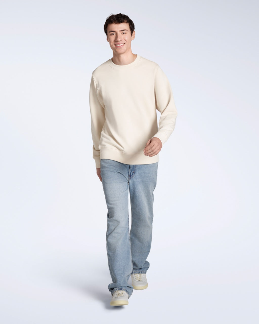 A full body photo of a man smiling walking towards the camera, modelling a plain natural coloured, organic cotton sweatshirt with a pair of blue jeans.
