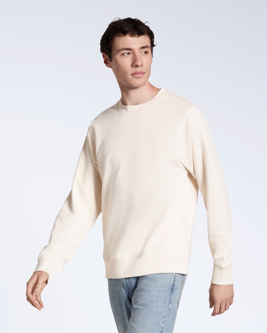 A man looking away from the camera, modelling a plain natural coloured, organic cotton sweatshirt with a pair of blue jeans.