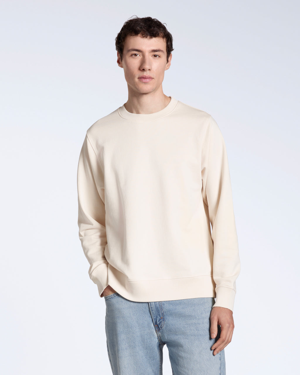 A man with his hand in his pocket looking into the camera, modelling a plain natural coloured, organic cotton sweatshirt with a pair of blue jeans.