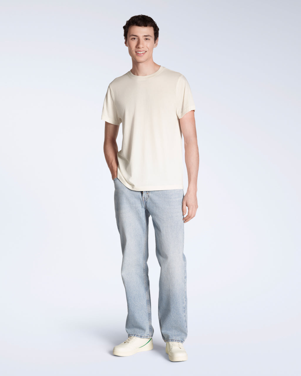 A full body photo of a man smiling into the camera with his hand in his pocket, modelling a plain natural coloured, short sleeve organic cotton t shirt with a pair of jeans.