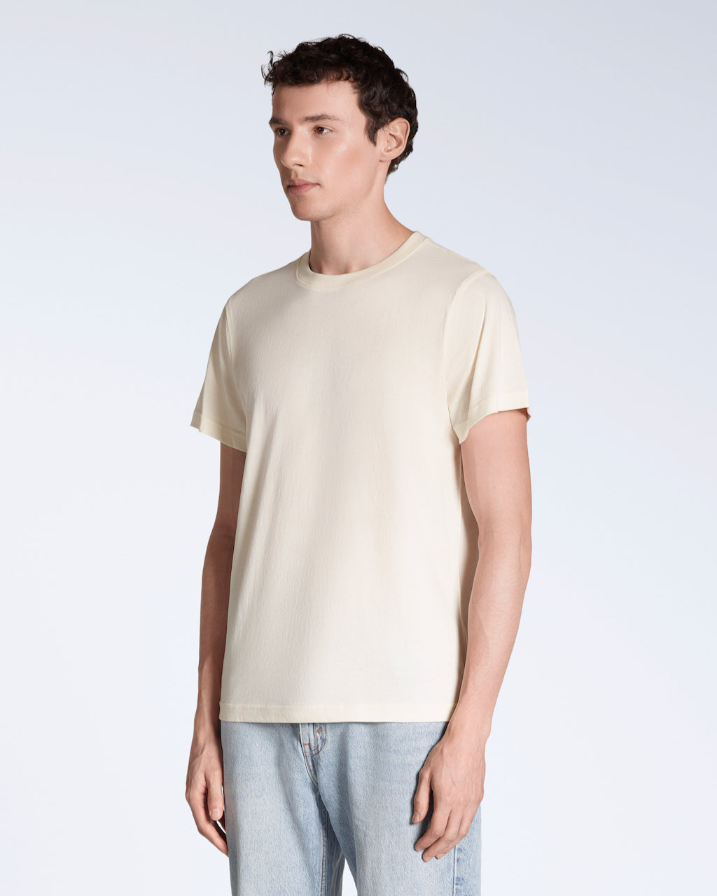 A man at an angle with his arms by his sides, modelling a plain natural coloured short sleeve organic cotton t shirt.