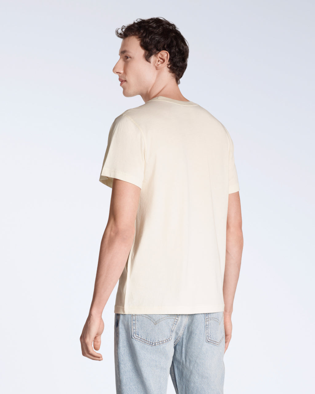 A man facing away from the camera, modelling a plain natural coloured, short sleeve, organic cotton t shirt.