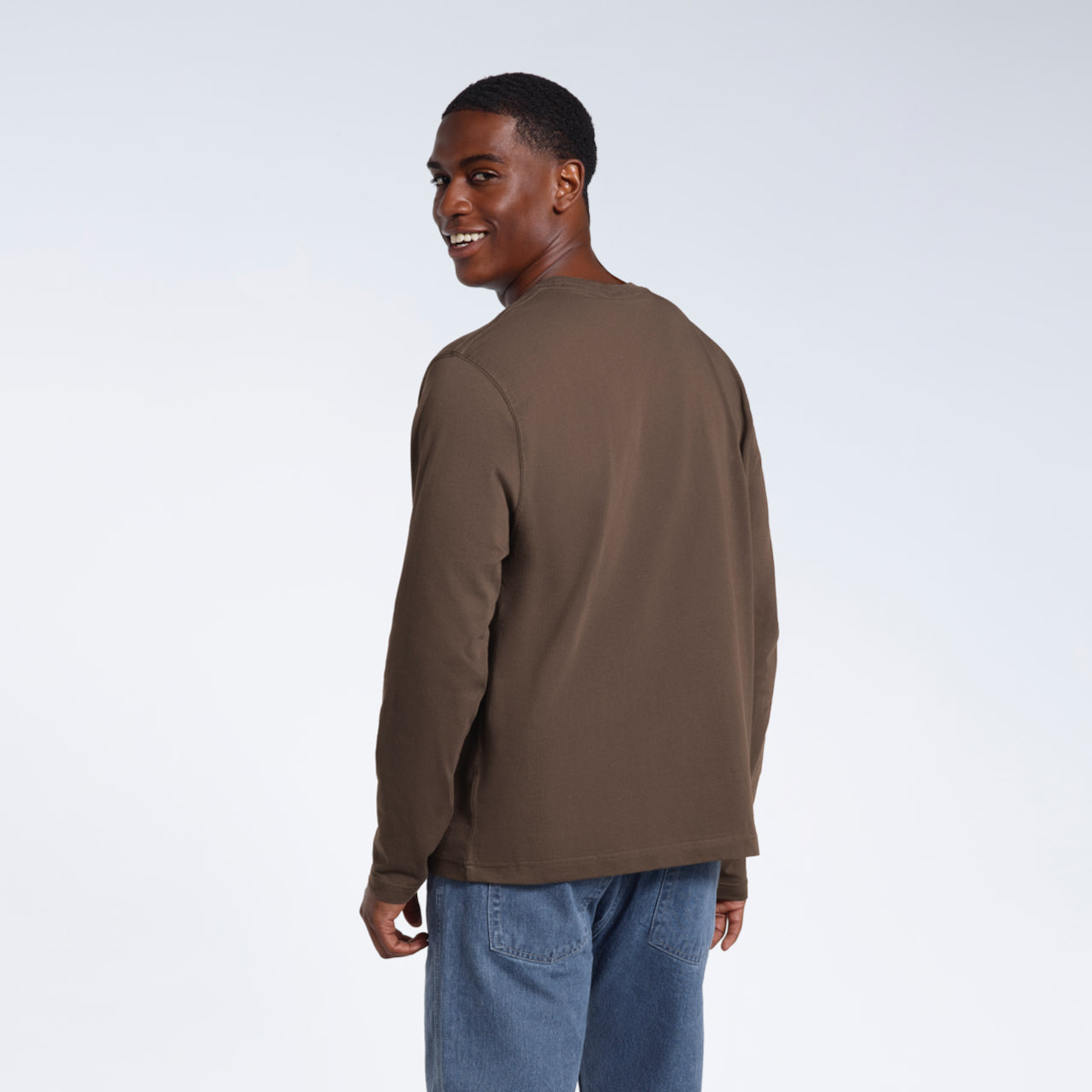 A man facing away from the camera looking back and smiling, modelling a plain dark khaki long sleeve organic cotton t shirt.