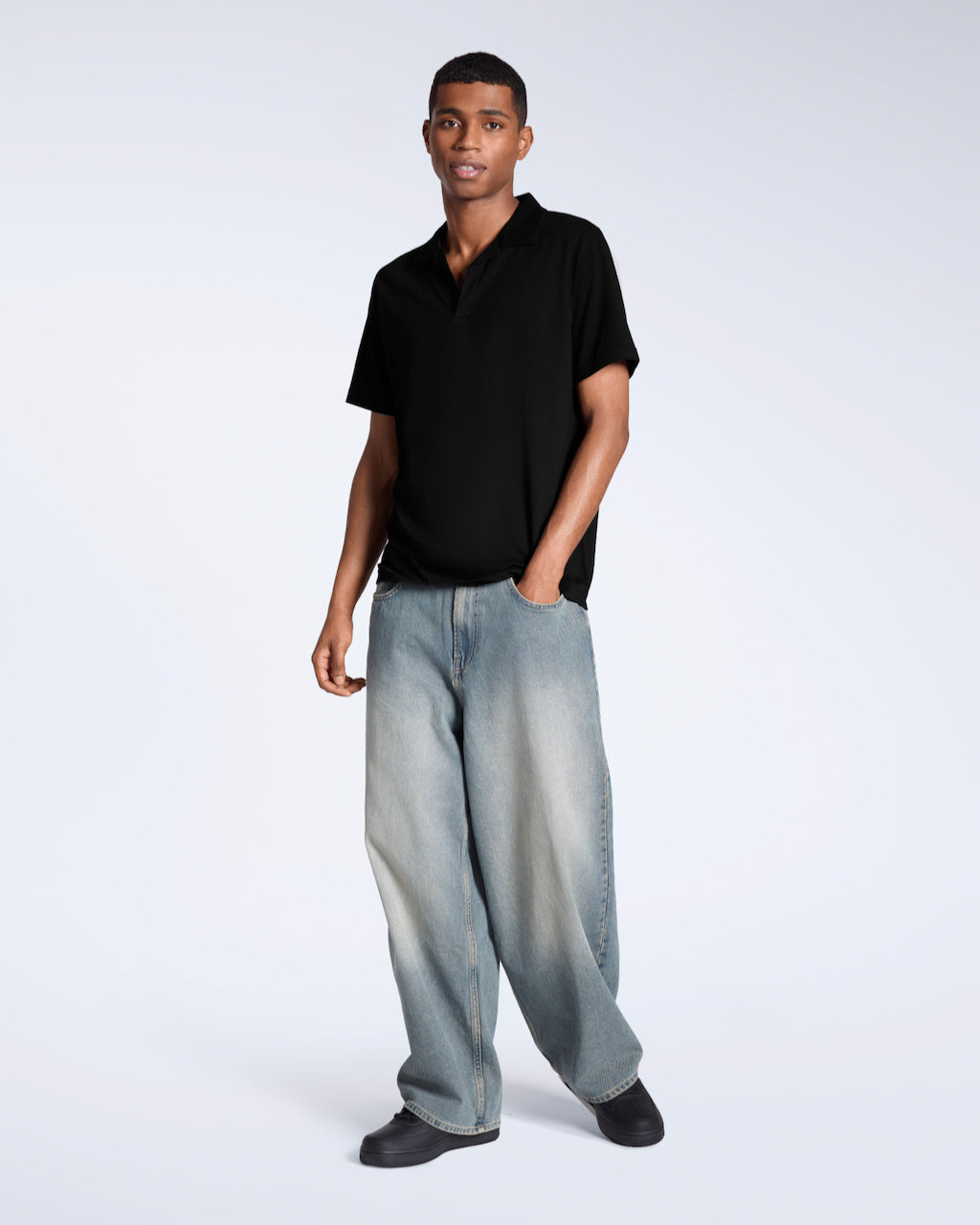 A full body photo of a man looking into the camera modelling a plain black, organic cotton, polo shirt with a pair of jeans.