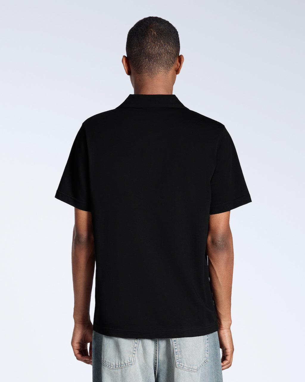A man facing away from the camera, modelling a plain black, short sleeve, organic cotton, polo shirt.