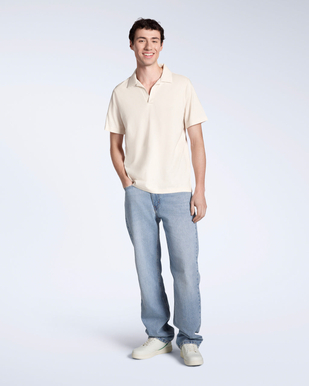 A full body photo of a man smiling into the camera with his hand in his pocket, modelling a plain, natural coloured, organic cotton polo shirt, with a pair of blue jeans.
