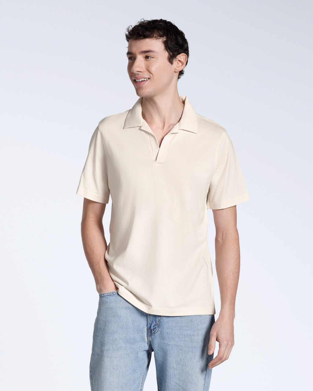 A man looking away from the camera with his hand in his pocket, modelling a plain natural coloured, short sleeve organic cotton polo shirt.