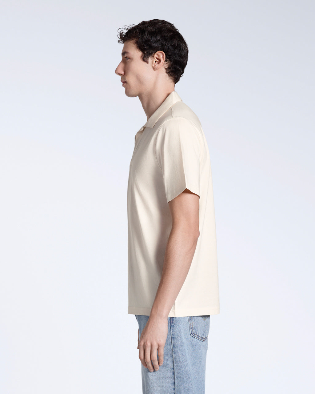 A man facing side on, modelling a plain natural coloured, short sleeve, organic cotton polo shirt.