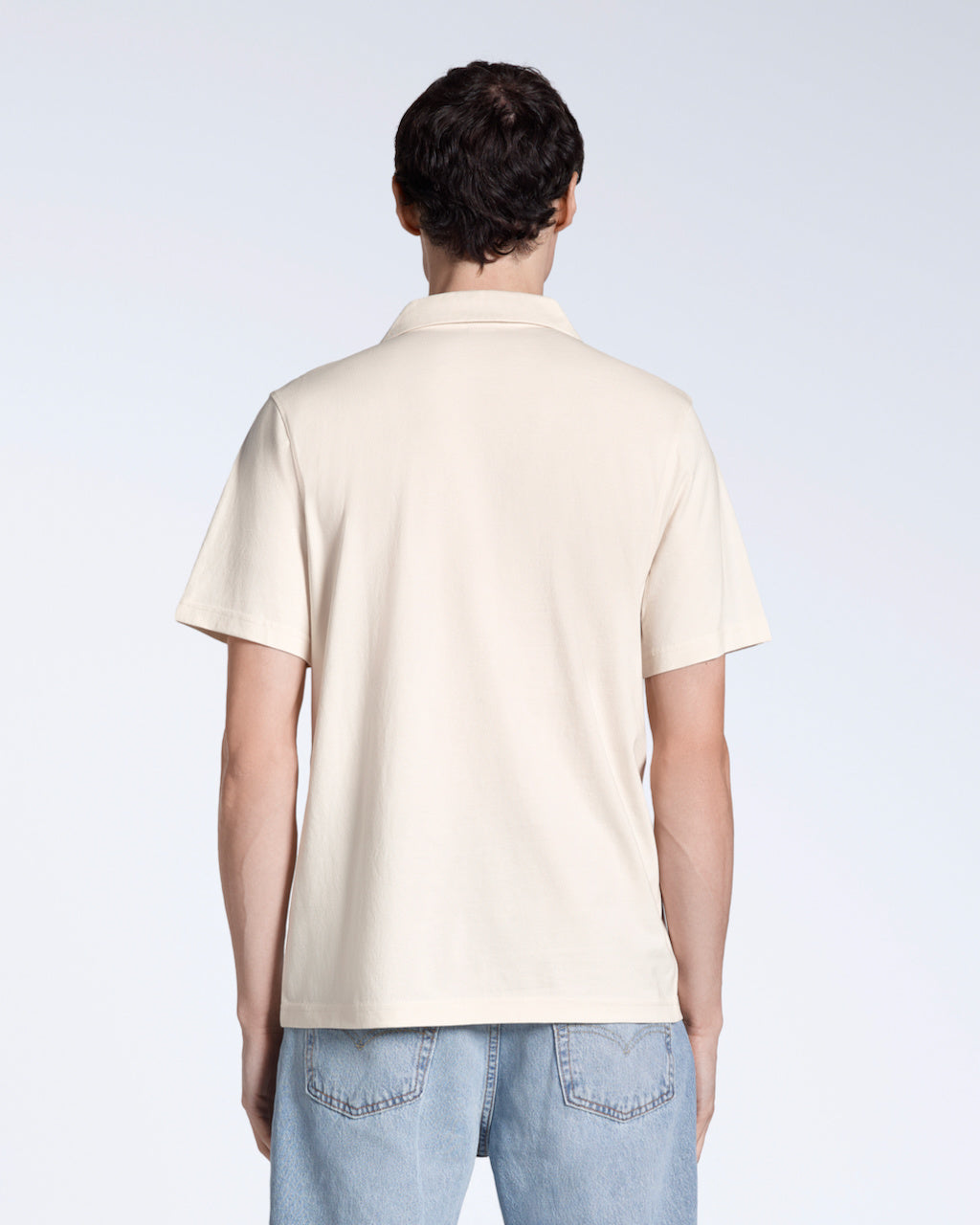 A man facing away from the camera, modelling a plain natural coloured, short sleeve, organic cotton, polo shirt.