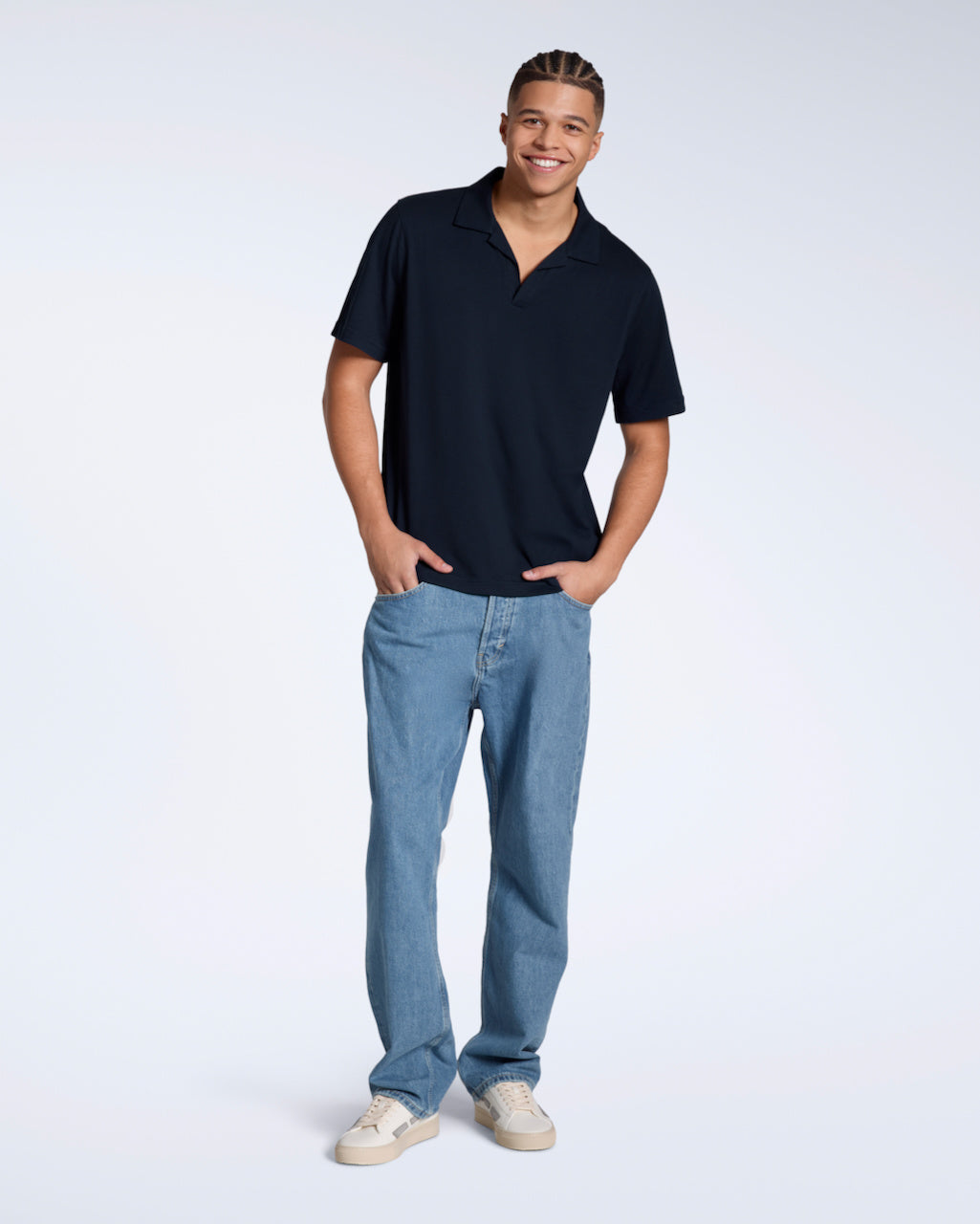 A full body photo of a man smiling into the camera with his hands in his pockets, modelling a plain navy, organic cotton, polo shirt with a pair of jeans.