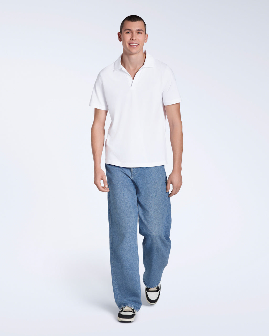A full body photo of a man smiling into the camera, modelling a plain white, organic cotton open neck polo shirt with a pair of jeans.