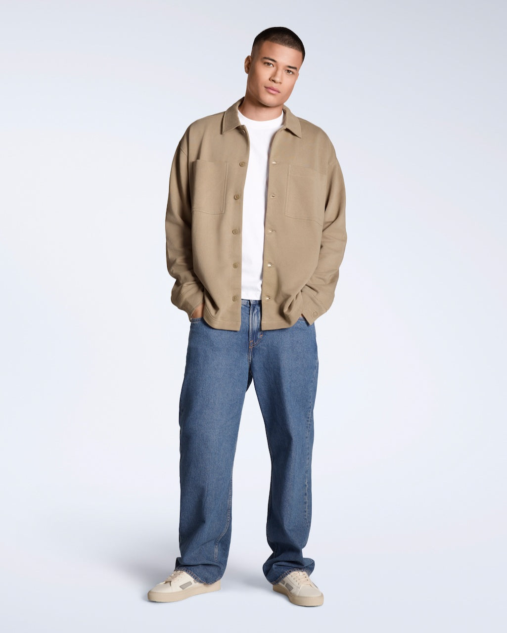A full body photo of a man looking into the camera with his hands in his pockets, modelling a plain beige, organic cotton overtshirt with a pair of jeans.
