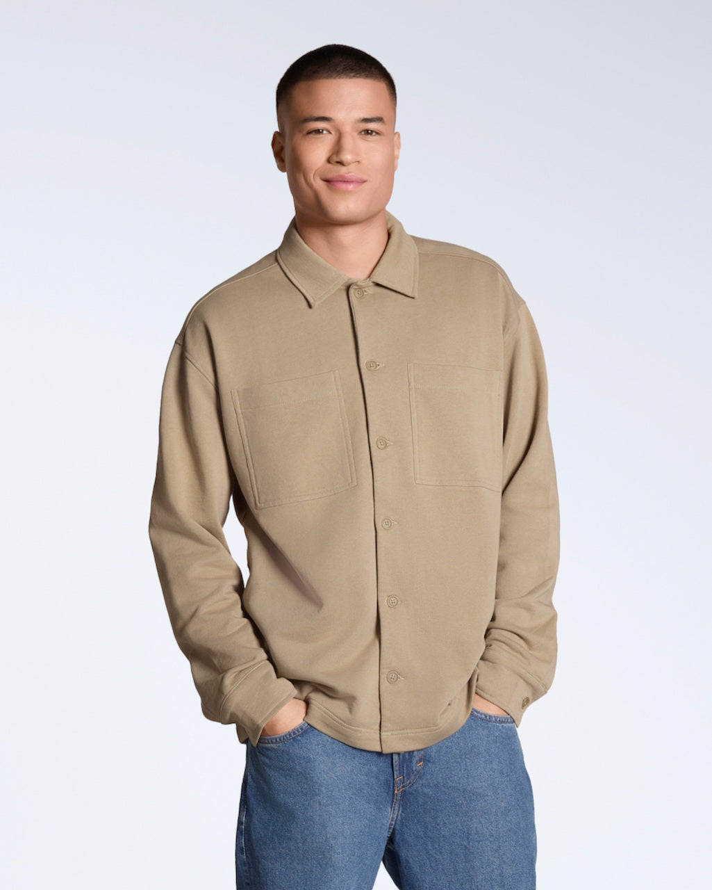 A man smiling into the camera with his hands in his pockets, modelling a plain beige, organic cotton overshirt.