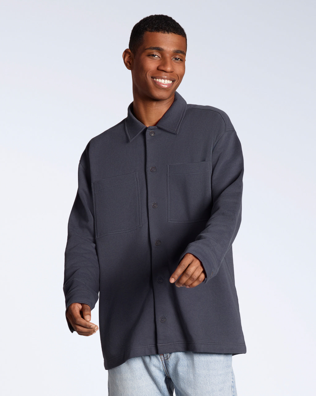 A man smiling into the camera, modelling a plain dark grey, organic cotton overshirt.