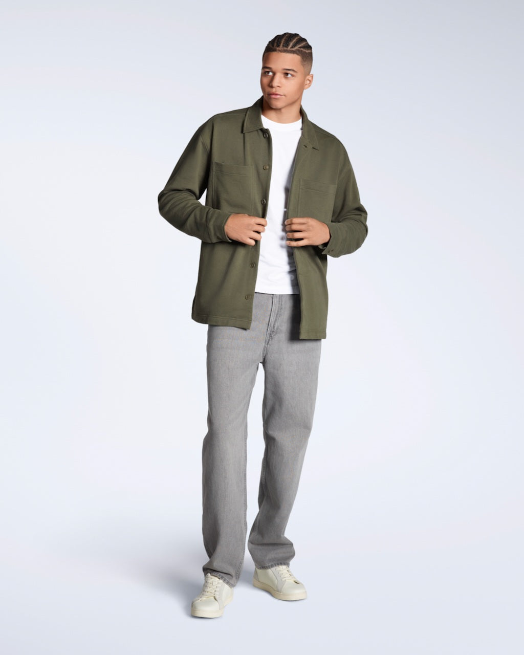 A full body photo of a man looking away from the camera, modelling a plain olive coloured, organic cotton overtshirt with a pair of jeans.