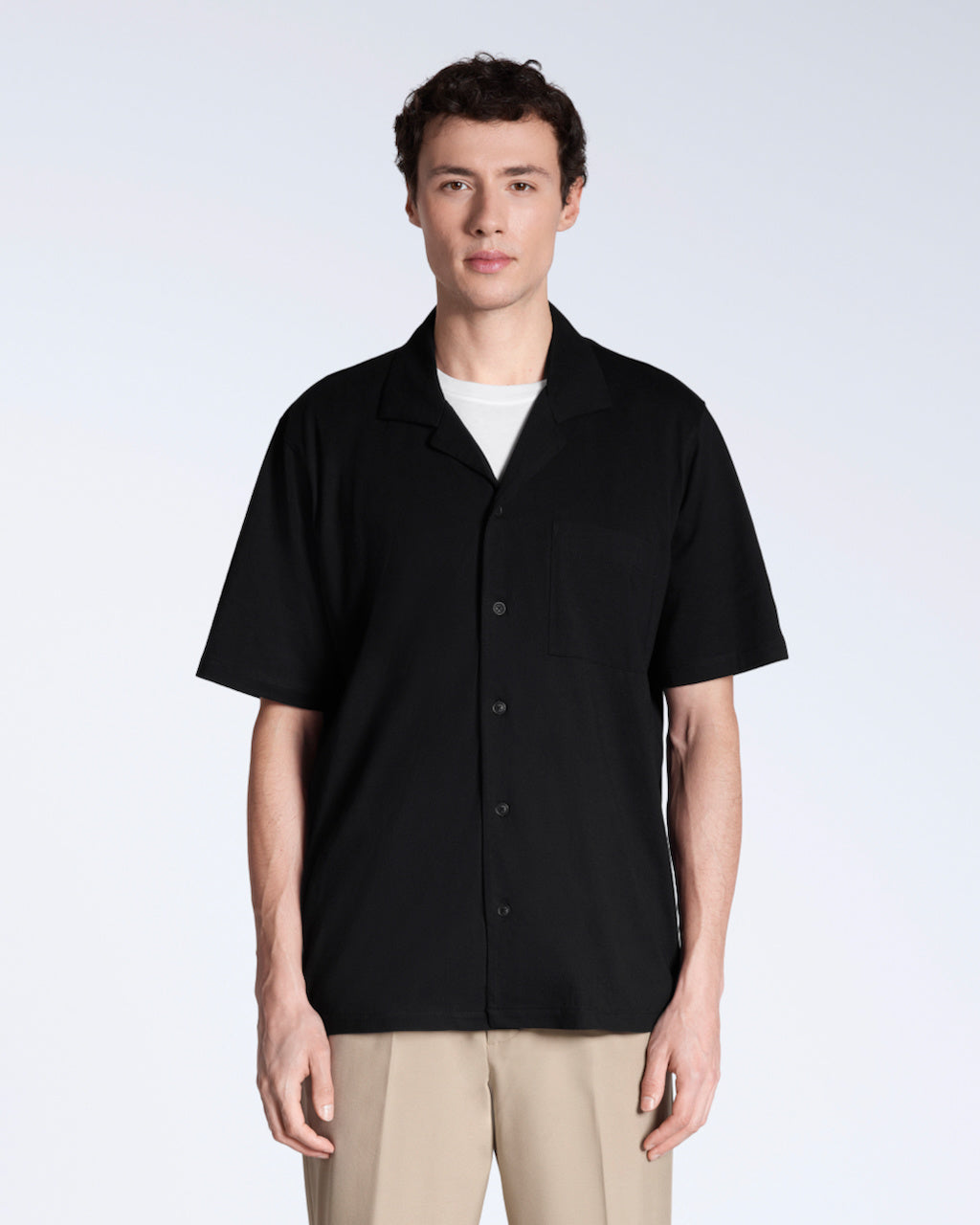 A man facing front on looking into the camera, modelling a plain black, organic cotton resort shirt.