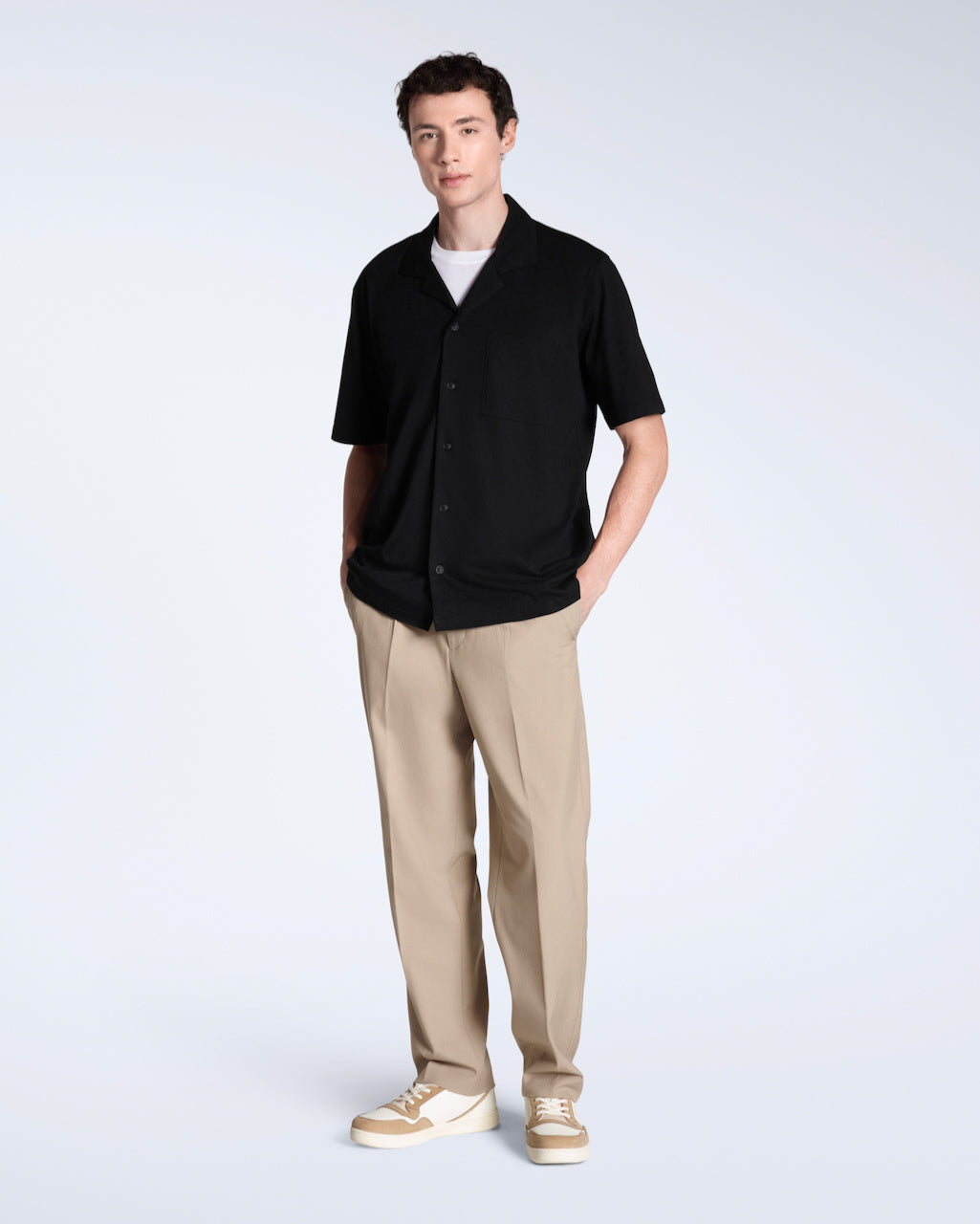 A full body photo of a man looking into the camera, modelling a plain black, organic cotton resort shirt with a pair of beige trousers.
