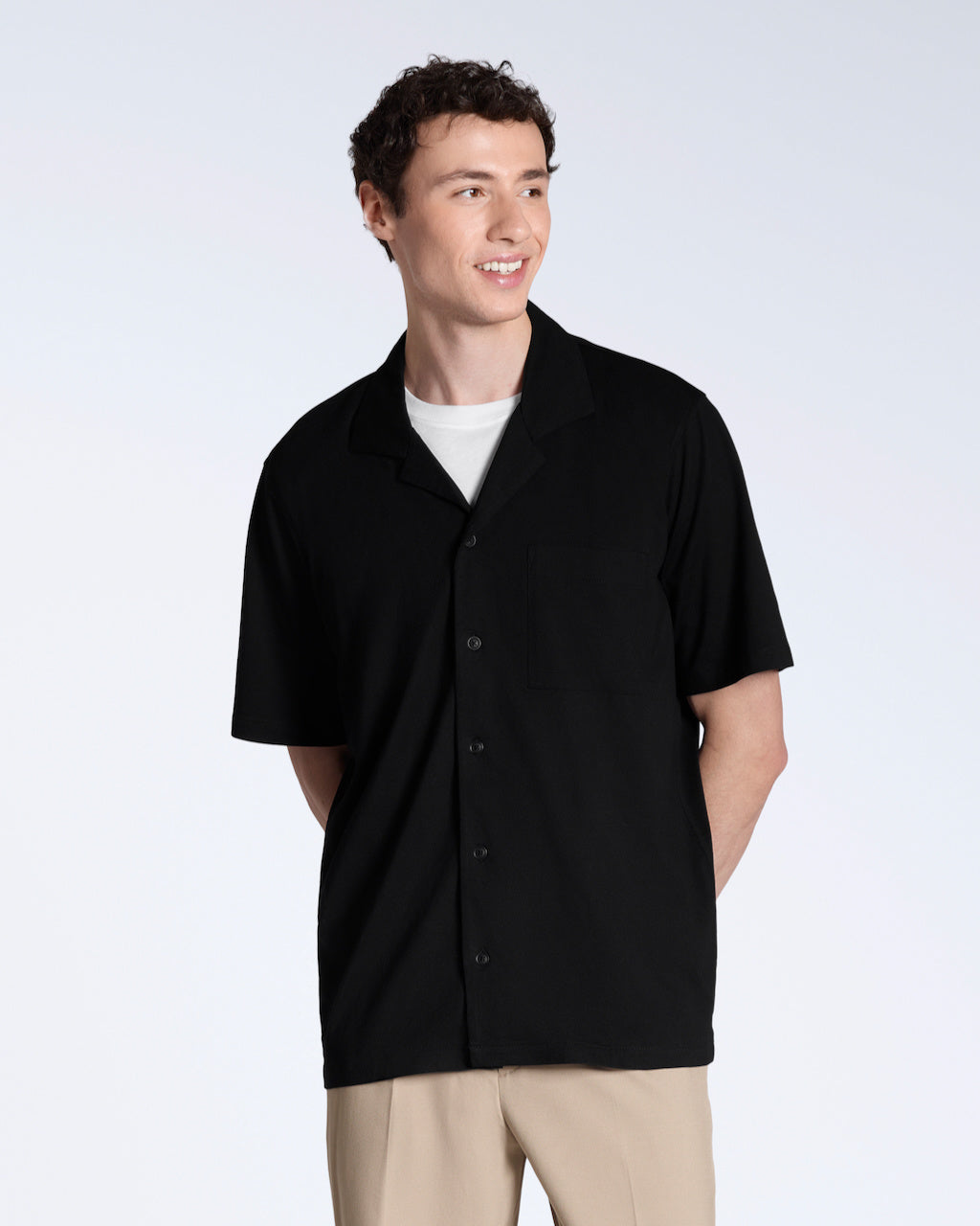 A man smiling, looking away from the camera, modelling a plain black, organic cotton resort shirt with a pair of beige trousers.