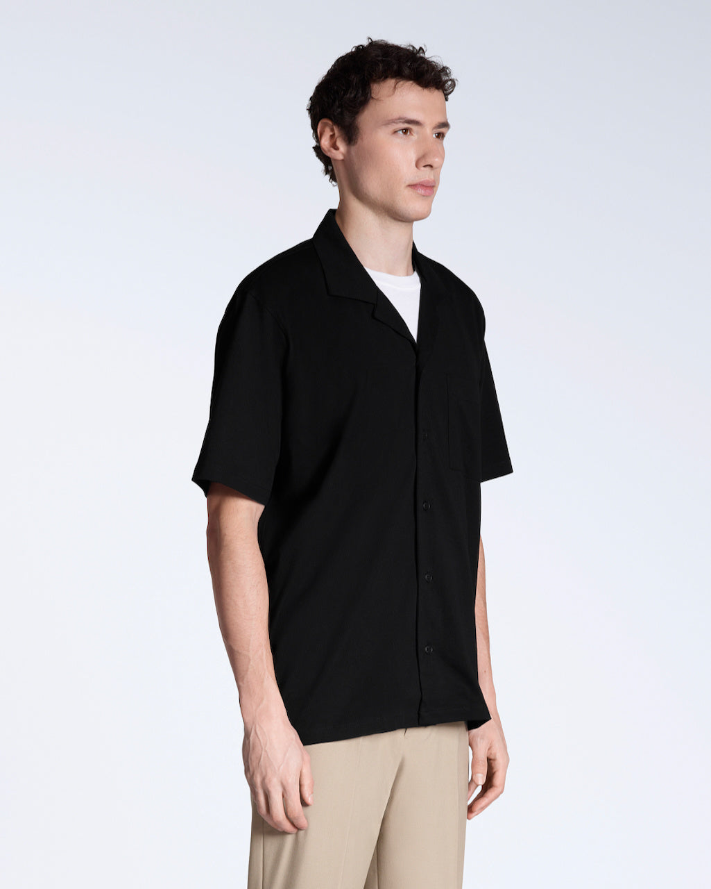 A man at an angle with his arms by his sides, modelling a plain black, organic cotton resort shirt.