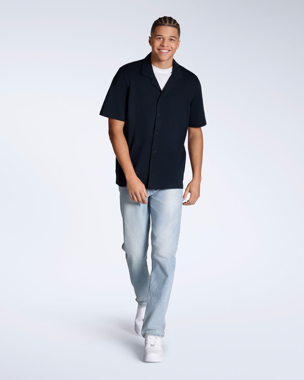 A full body photo of a man smiling into the camera, modelling a plain navy, organic cotton resort shirt with a pair of jeans.
