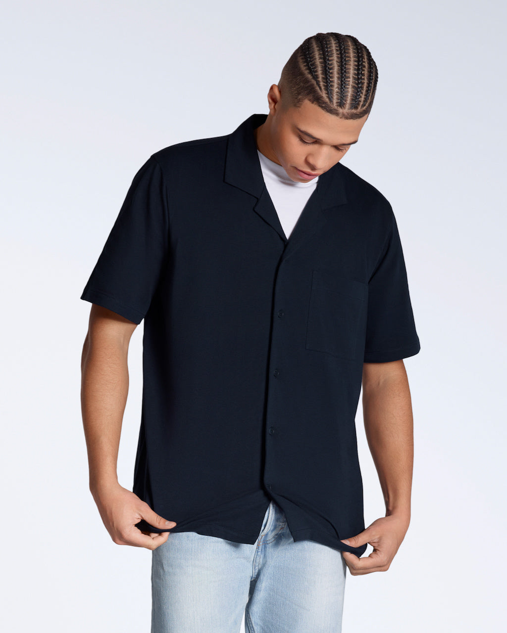 A man looking down touching the bottom of his shirt, modelling a plain navy, organic cotton resort shirt.
