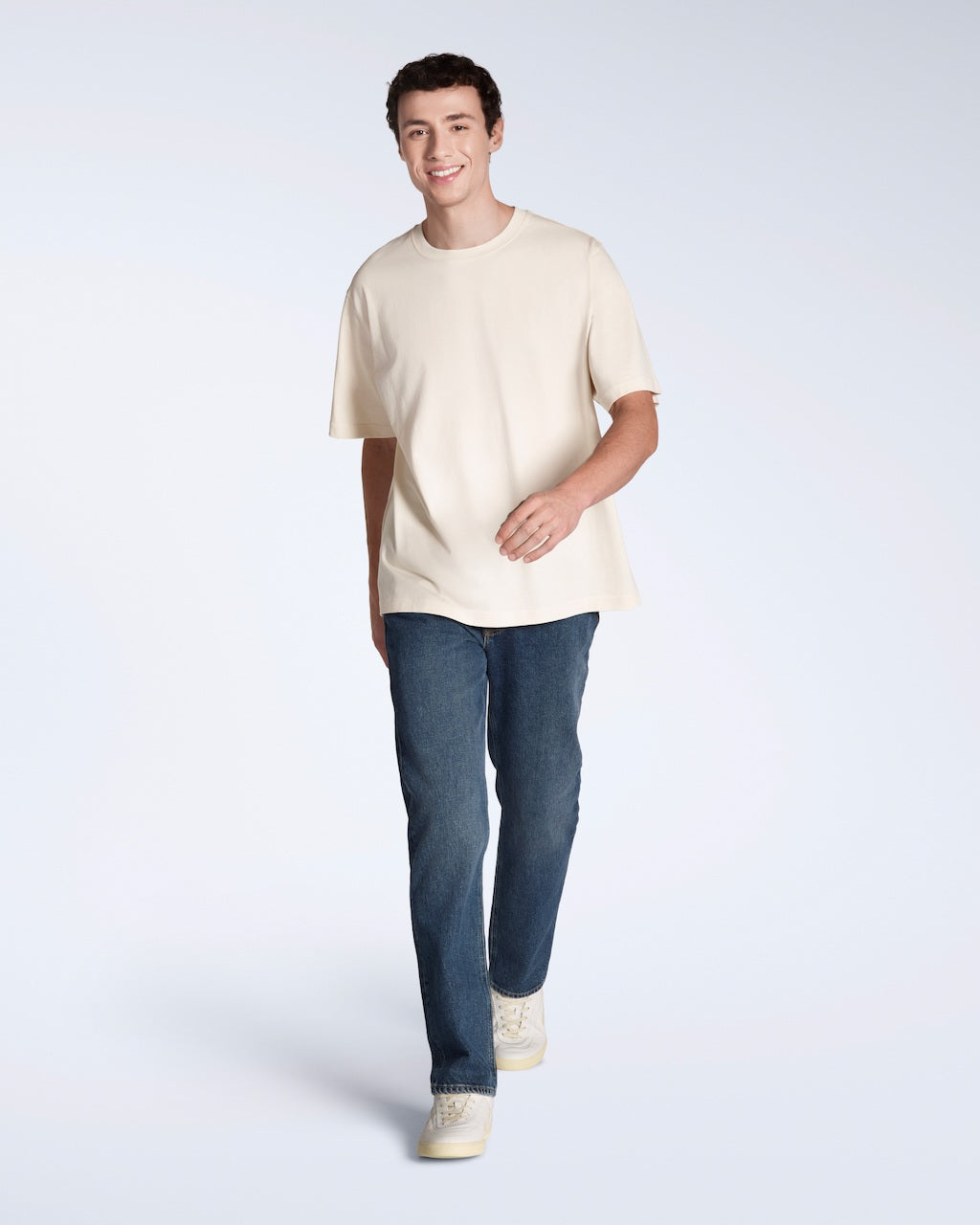 A full body photo of a man smiling, walking towards the camera, modelling a plain natural coloured, oversized, organic cotton t shirt.