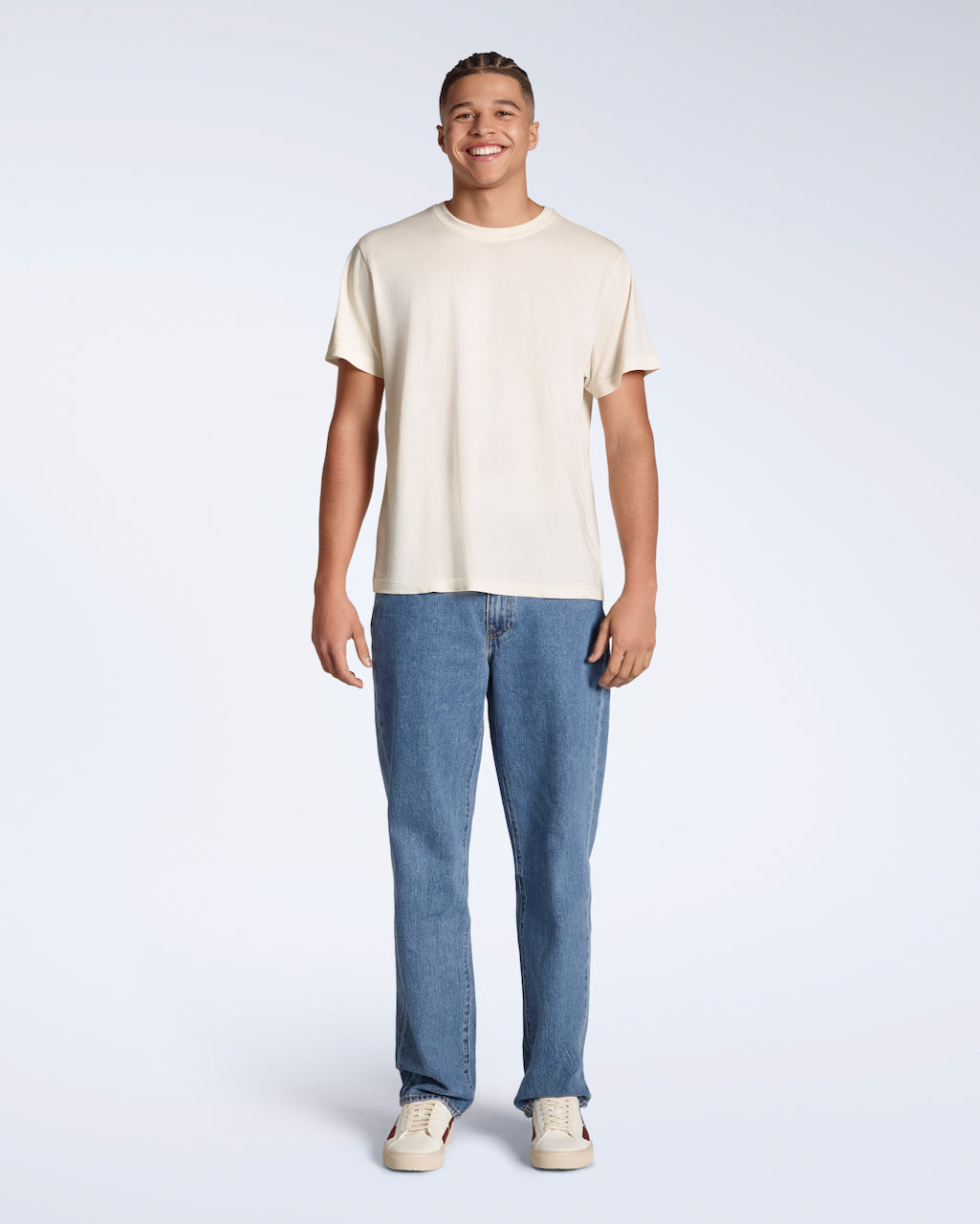 A full body photo of a man smiling into the camera, modelling a plain, natural coloured, short sleeve organic cotton t shirt with a pair of jeans.