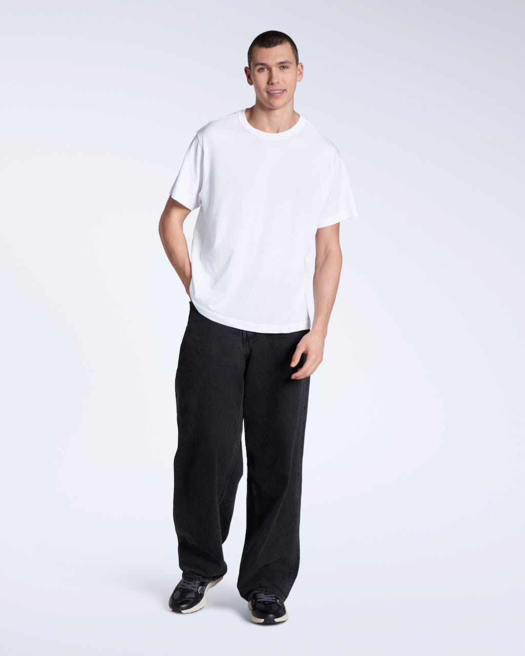 A man smiing into the camera with a hand in his pocket, modelling a plain white, organic cotton t shirt with a pair of black jeans.