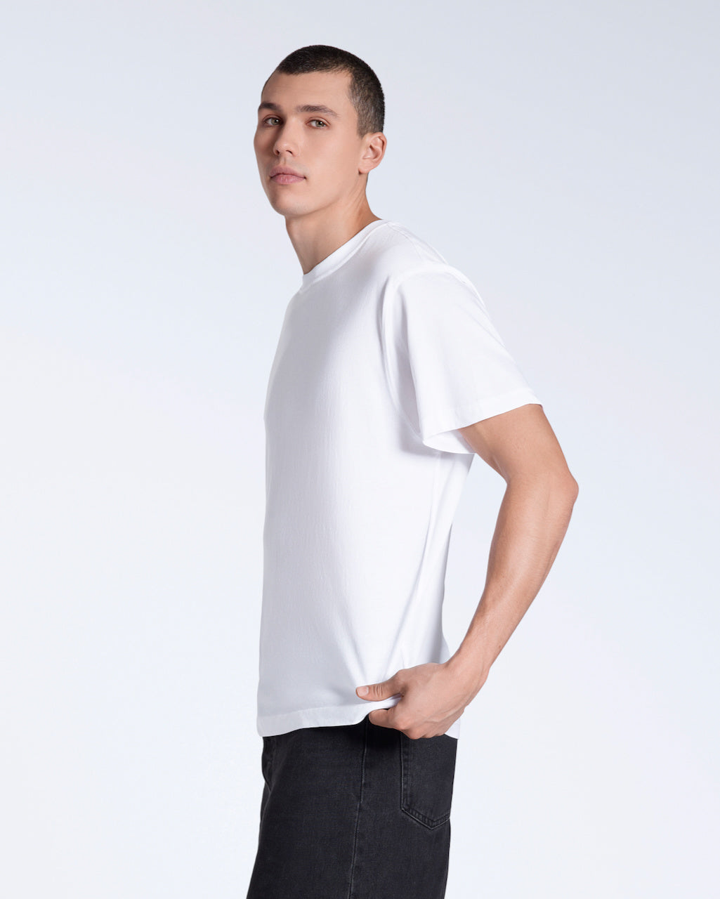 A man facing side on looking into the camera, modelling a plain white, short sleeve organic cotton t shirt.