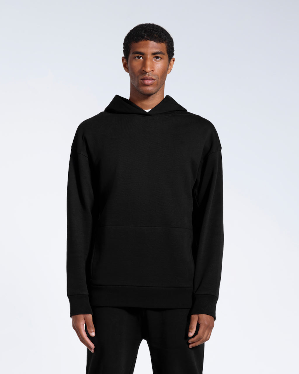 A man facing front on looking into the camera with his arms by his sides, modelling a black oversized organic cotton hoodie.