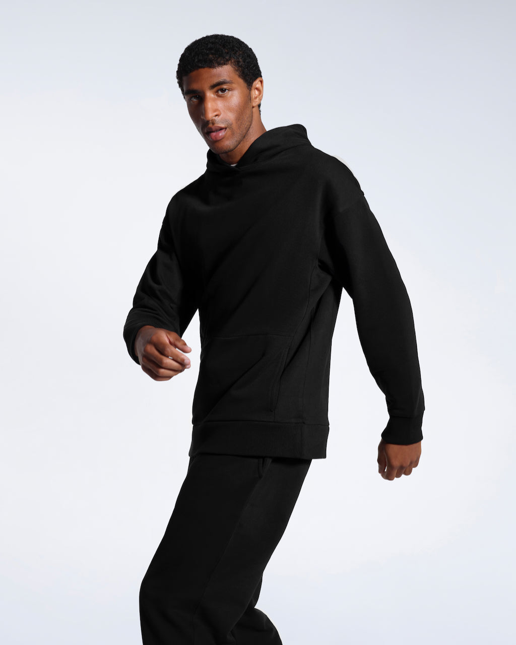 A man mid movement looking into the camera, modelling a black oversized organic cotton hoodie with matching joggers.