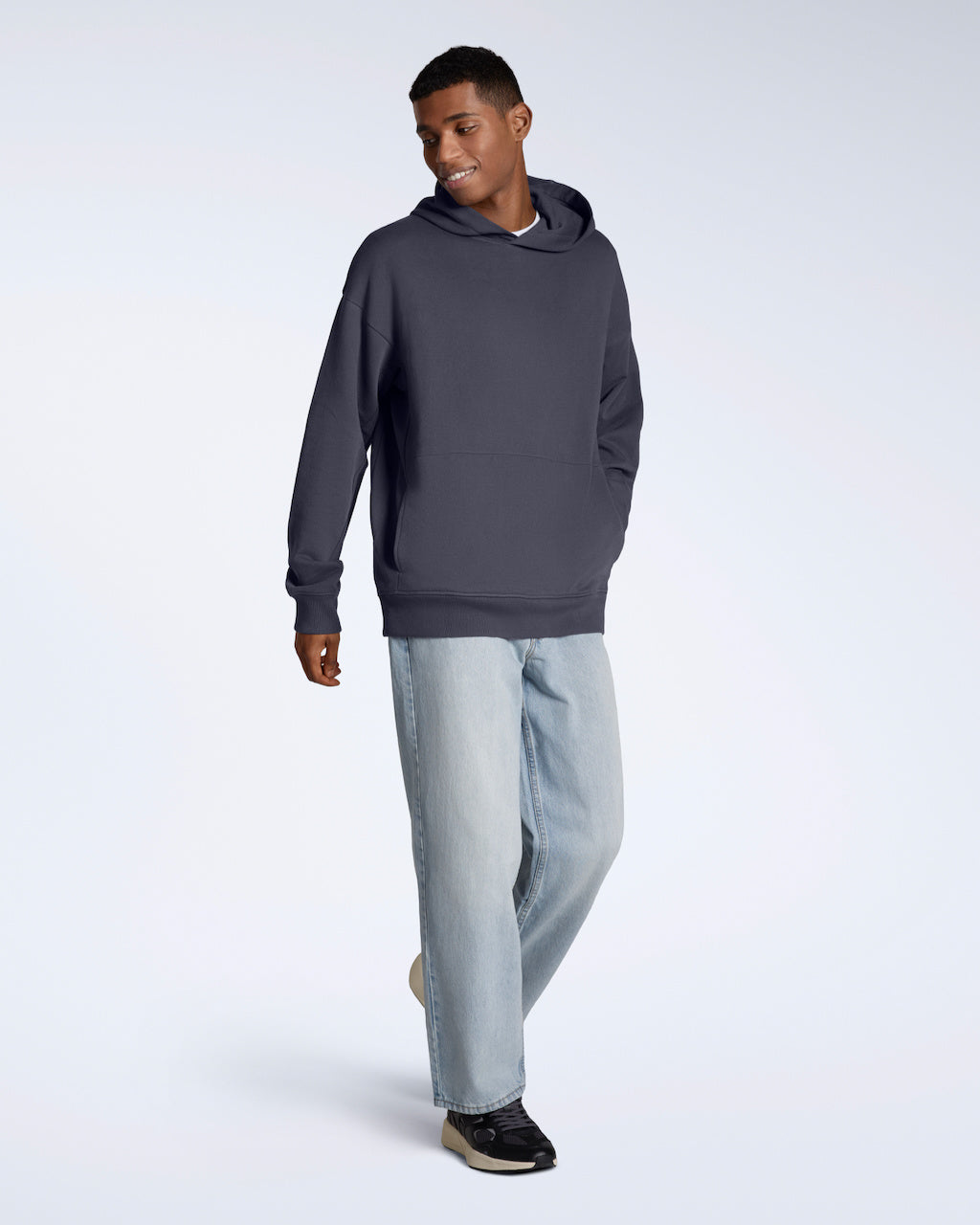 A full body photo of a man walking and smiling, modelling a dark grey oversized organic cotton hoodie with a pair of jeans.