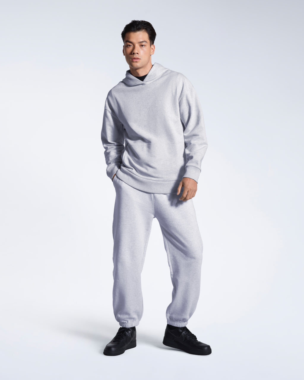 A full body photo of a man looking into the camera with a hand in his pocket, modelling a grey oversized organic cotton hoodie with matching joggers.