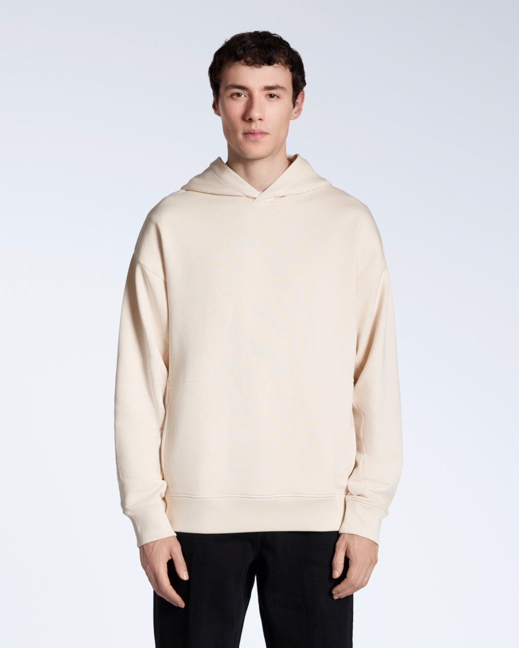 A man facing front on looking into the camera with his arms by his sides, modelling a natural coloured oversized organic cotton hoodie.