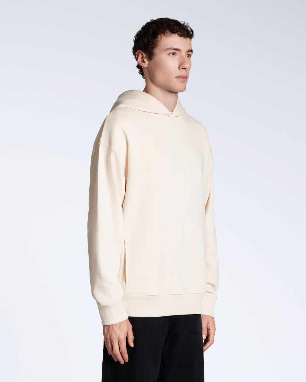 A man at an angle looking away from the camera with his arms by his side, modelling a natural coloured oversized organic cotton hoodie.