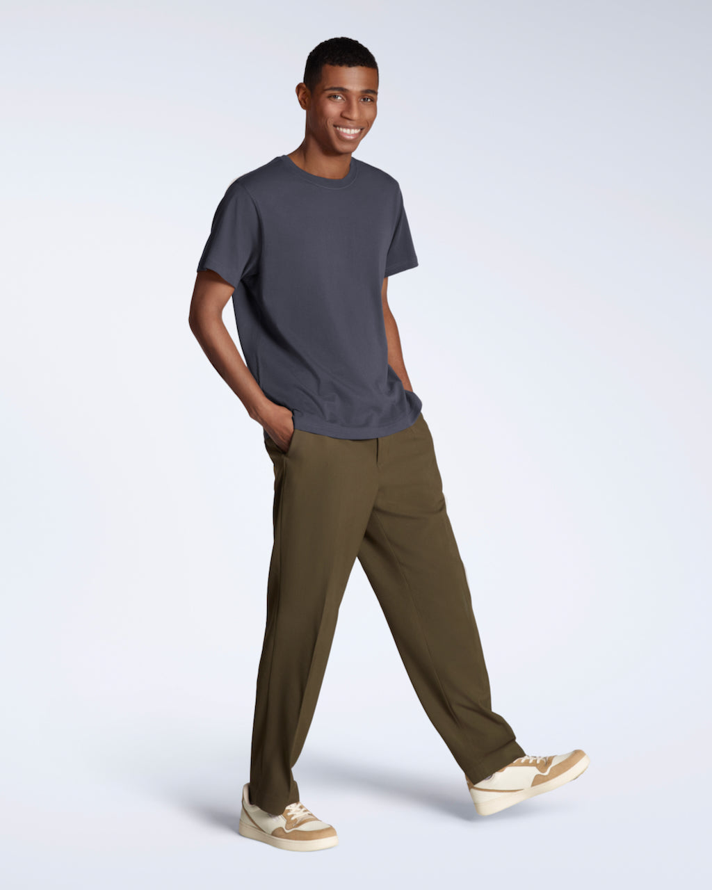 A full body photo of a man smiling into the camera with his hands in his pockets, modelling a plain dark grey, short sleeve organic cotton t shirt with a pair of trousers.