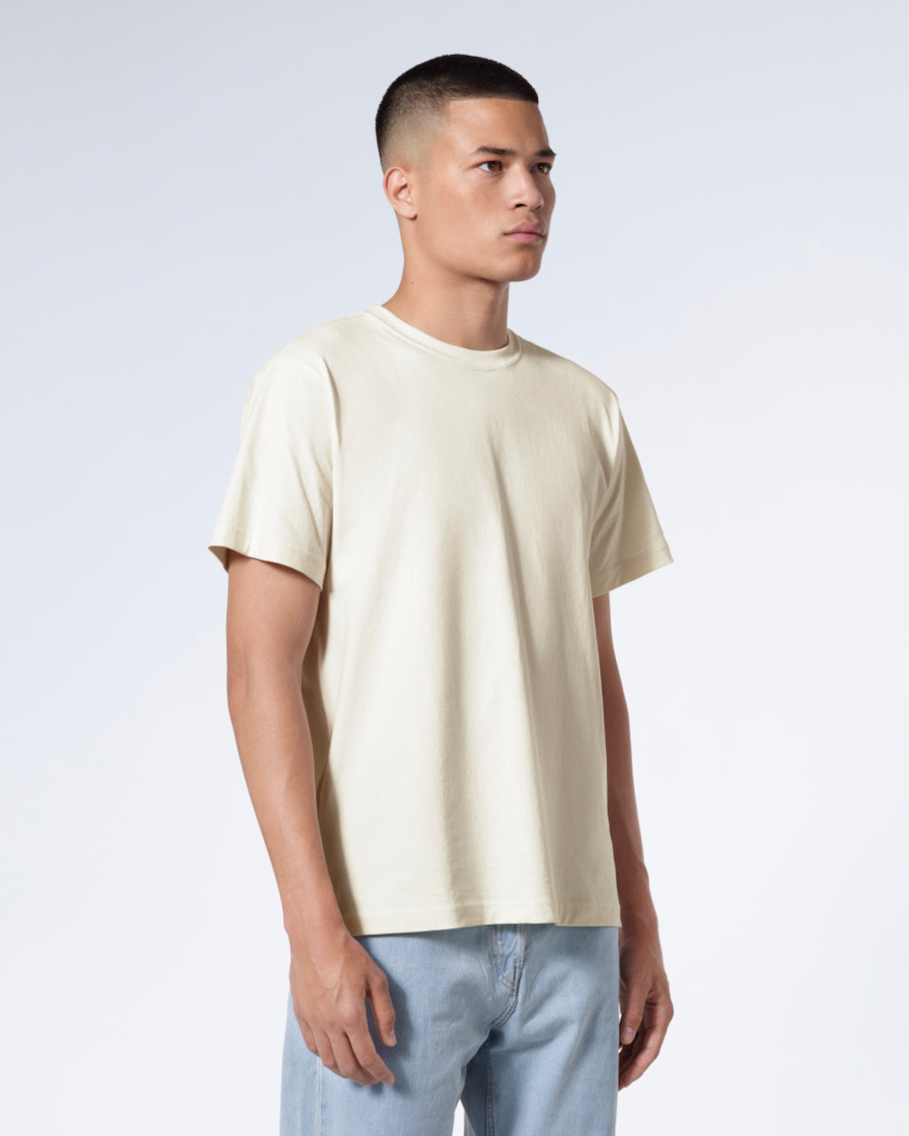 A man at an angle with his arms by his sides, modelling a plain ecru short sleeve organic cotton t shirt.