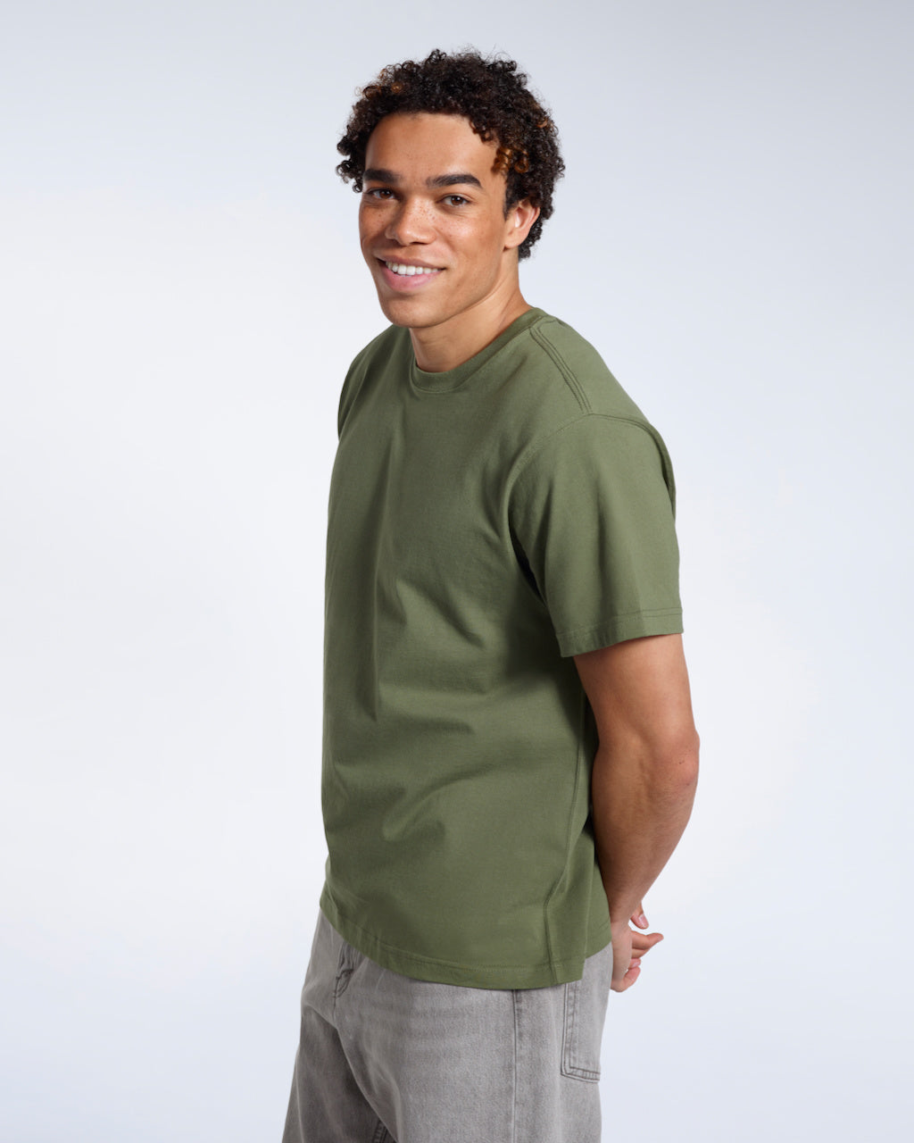 A man smiling into the camera with his hands behind his back, modelling a plain khaki green short sleeve organic cotton t shirt.