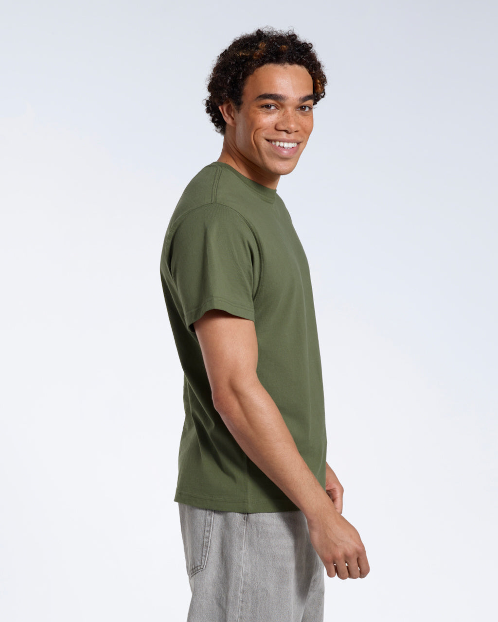 A man facing side on with his head turned towards the camera smiling, modelling a plain khaki green short sleeve organic cotton t shirt.